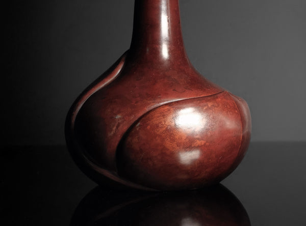 A glossy reddish-brown ceramic vase with a bulbous rounded body and a narrow neck, photographed on a dark reflective surface against a dark gray background.