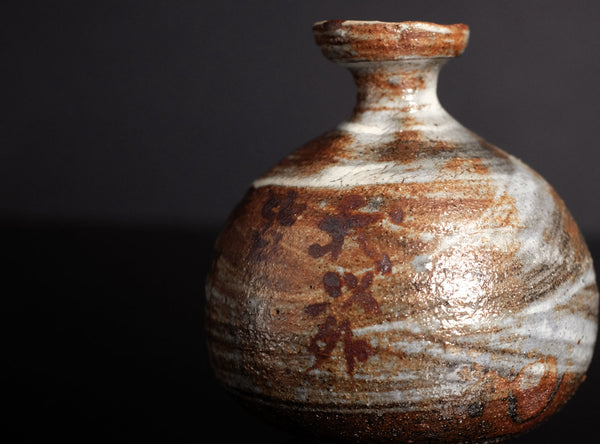 A close-up of a handmade ceramic bottle with a rounded body, narrow neck, and flared rim, featuring a textured glaze in earthy brown, white, and black marbled patterns against a dark background.