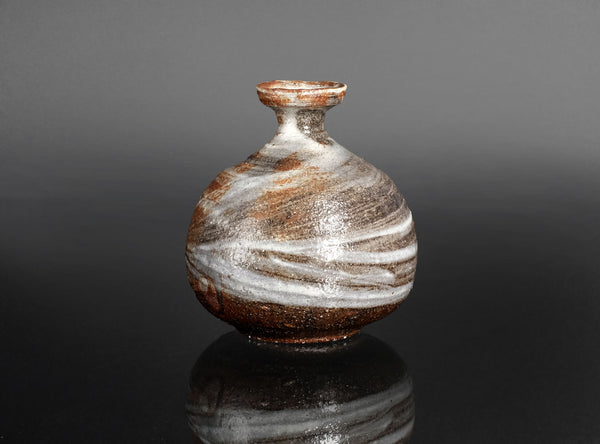 A small round ceramic vase with a narrow neck, featuring a marbled glaze of brown, white, and gray, displayed on a reflective black surface against a dark gray background.