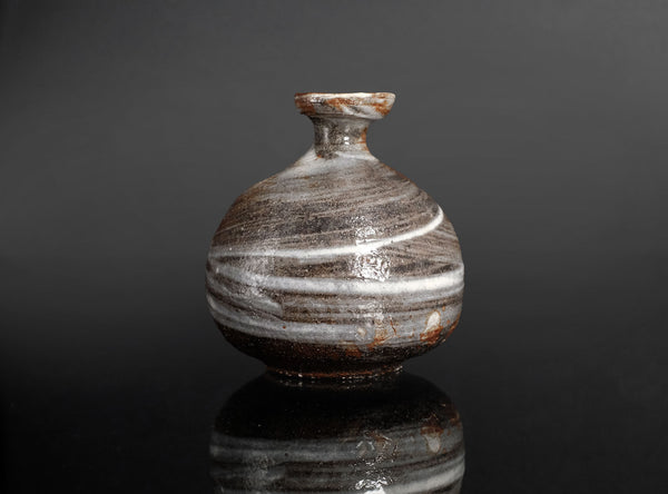 A small round ceramic vase with a narrow neck and flared rim, featuring horizontal bands of glossy brown, gray, and white glaze, set against a dark reflective background.