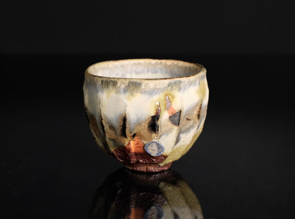 A handmade ceramic tea cup with a textured, marbled glaze in cream, brown, black, and subtle copper-orange tones, set against a solid black background with a soft reflection below.