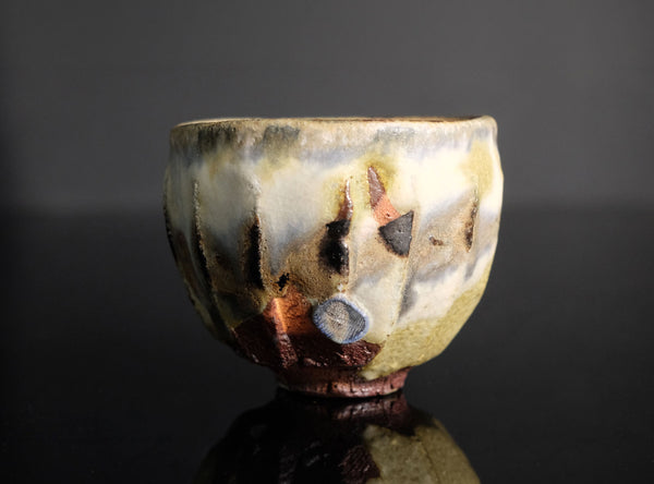 An abstract ceramic cup with a textured, organic glaze in cream, brown, black, and muted copper tones, set against a dark reflective background.