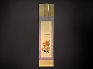 A traditional Japanese hanging scroll (kakejiku) featuring a painted samurai warrior in red and gold armor, framed by purple patterned borders and gold-toned paper bands at the top and bottom, displayed against a dark background.