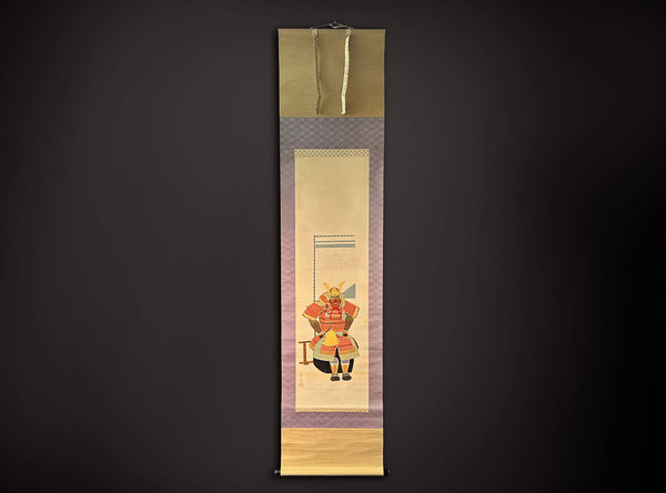 A traditional Japanese hanging scroll (kakejiku) featuring a painted samurai warrior in red and gold armor, framed by purple patterned borders and gold-toned paper bands at the top and bottom, displayed against a dark background.