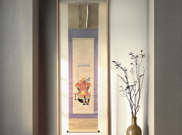 A vertical Japanese-style hanging scroll artwork displayed in a narrow frame, featuring a samurai warrior illustration. The scroll hangs against a light wall next to a vase with dried branches.