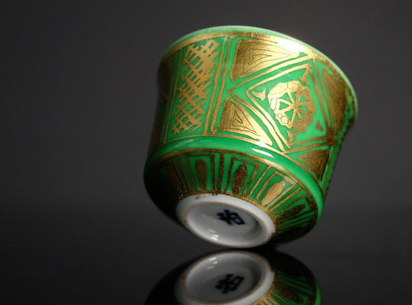 Decorative ceramic cup with green and gold patterns on a reflective surface.