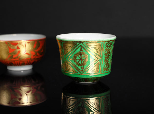 Two ornate cups, one red and gold, the other green and gold, on a reflective black surface.