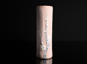 A tall, cylindrical ceramic vase with a textured, crackled glaze in pale pink and white, standing upright on a reflective black surface against a solid black background.