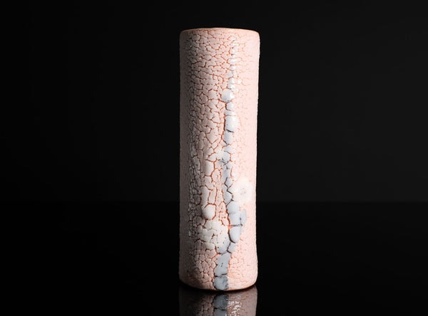 A tall, cylindrical ceramic vase with a textured, crackled glaze in pale pink and white, standing upright on a reflective black surface against a solid black background.