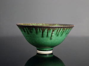 A green glazed ceramic bowl with a dark metallic drip pattern around the rim, set against a dark gray background with a reflective surface below.