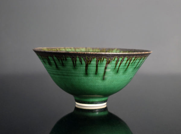 A green glazed ceramic bowl with a dark metallic drip pattern around the rim, set against a dark gray background with a reflective surface below.