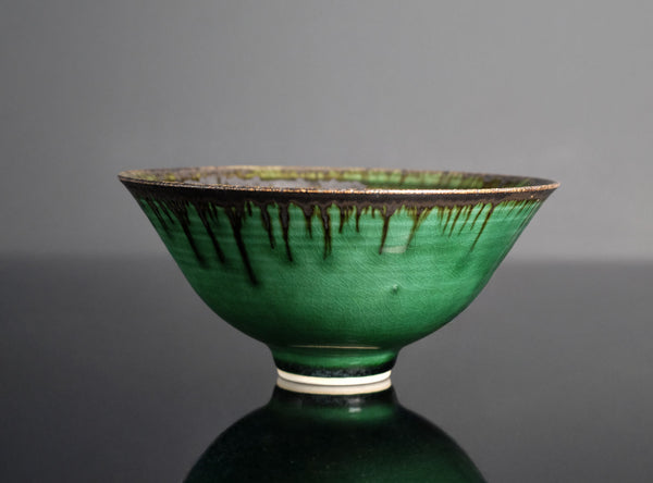A green glazed ceramic bowl with dark brown dripping glaze details and a white band near the base, set against a dark gray background.