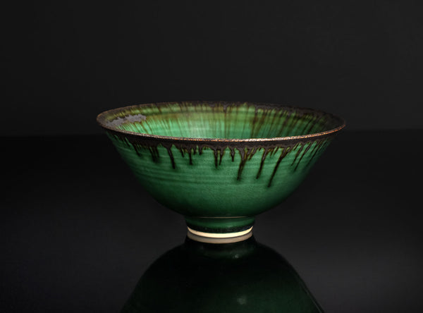 A green glazed ceramic bowl with dark dripping glaze details, displayed on a reflective black surface against a dark background.