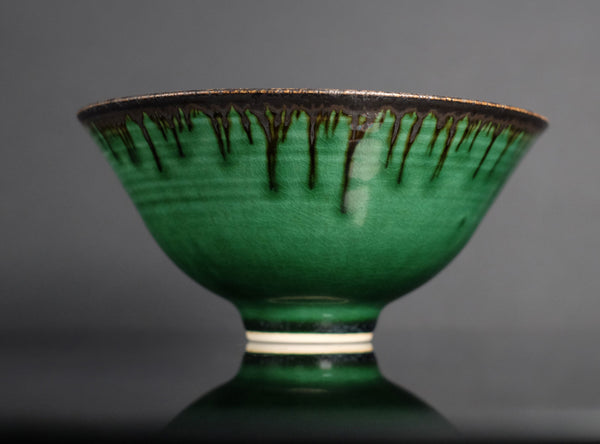 A green glazed ceramic bowl with a dark brown dripping glaze pattern around the rim and a small white band near the base, shown against a dark gray background.
