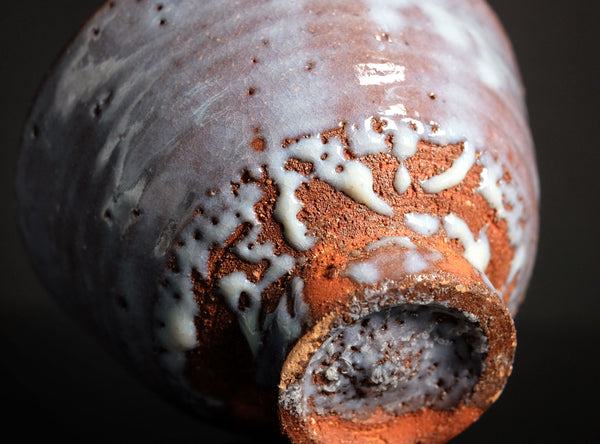 Close-up of a ceramic bowl with a glossy glaze showing a mottled blend of dark gray, warm brown, and white drips, with the open base visible against a dark background.