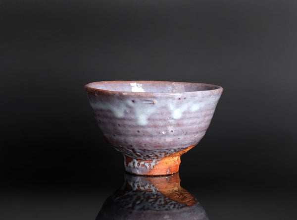 A handmade ceramic tea bowl with a textured, speckled glaze in earthy brown, white, and muted pink tones, shown on a reflective black surface against a dark gray background.