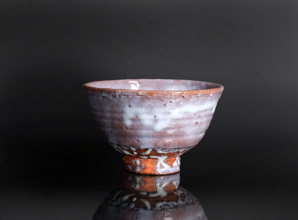 A handmade ceramic tea bowl with a textured, speckled glaze in earthy brown, white, and muted pink tones, shown on a reflective black surface against a dark gray background.