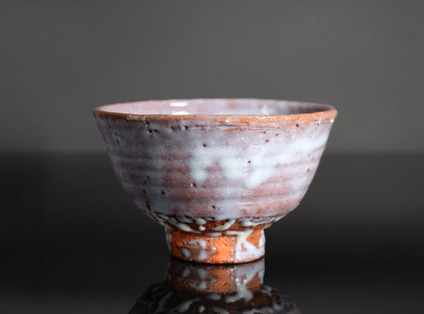 A handmade ceramic tea bowl with a textured, speckled glaze in earthy brown, white, and muted pink tones, shown on a reflective black surface against a dark gray background.