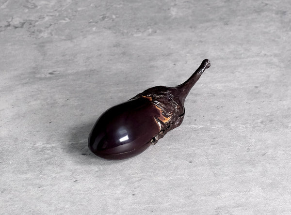 A glossy dark purple eggplant-shaped object with a long stem, photographed on a light gray textured surface.