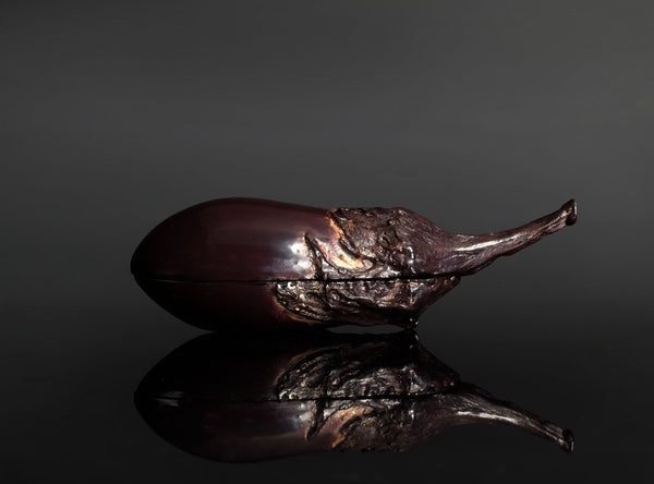 A sculptural eggplant-shaped object with a glossy dark purple body and textured brown stem, photographed on a reflective black surface against a dark gray background.