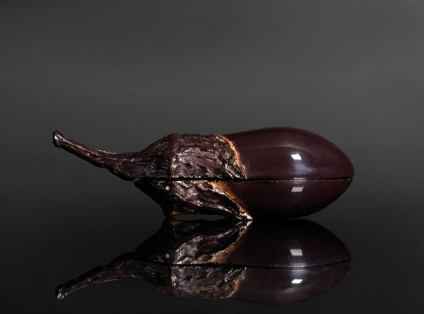A sculptural eggplant-shaped object with a glossy dark purple body and textured brown stem, photographed on a reflective black surface against a dark gray background.