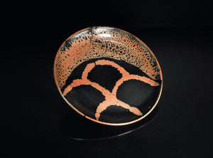 A round ceramic plate with a glossy black glaze, featuring a textured orange-brown speckled pattern around the rim and a large abstract orange-brown geometric motif on the bottom half, set against a solid black background.