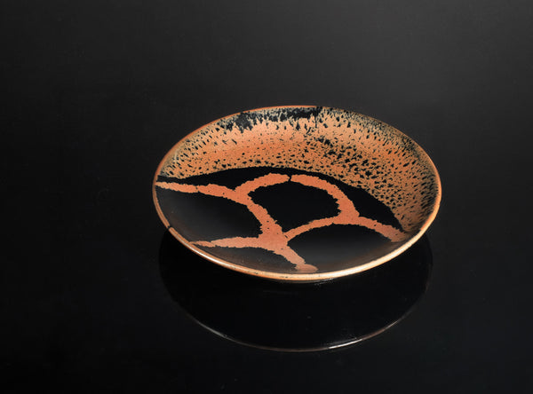 A round ceramic plate with a glossy black glaze, featuring a copper-orange branch-like pattern and fine speckling on the interior, set against a solid black background.