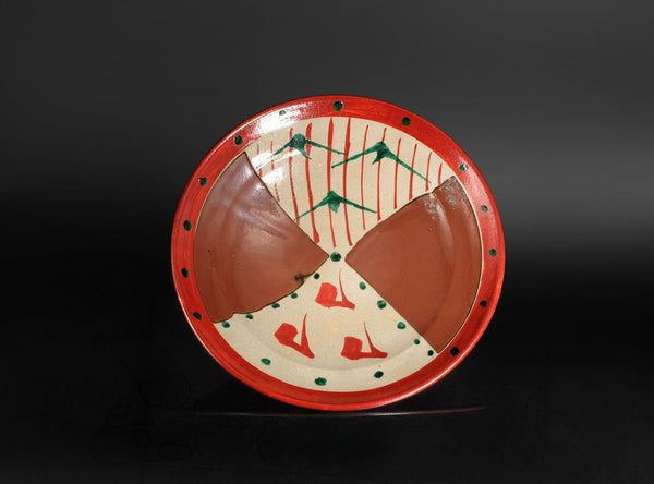 A round hand-painted ceramic plate with a red rim, featuring four geometric sections in terracotta, cream, red, and green, set against a dark background.