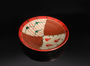A ceramic bowl with a split design, featuring a red outer rim, one half with red diagonal stripes on an off-white background, and the other half with terracotta-colored segments. The bottom half also has small red and green fish-like motifs on an off-white background, set against a dark reflective background.