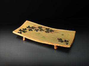 A rectangular ceramic serving tray with a light beige painted surface, decorated with black leaf motifs and small red berry accents, shown on a dark studio background.