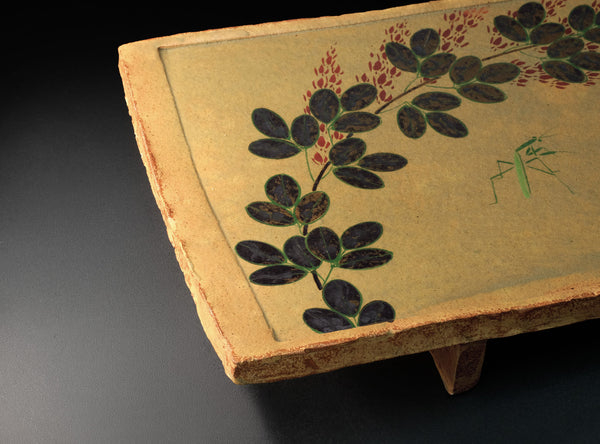 A rectangular ceramic serving tray with a light beige painted surface, decorated with black leaf motifs and small red berry accents, shown on a dark studio background.