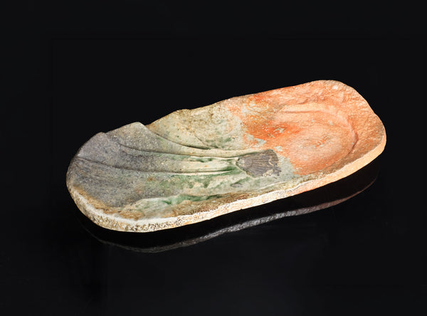 A rustic, hand-carved stone dish with a natural organic shape, featuring a mottled pattern of orange, green, and gray, displayed on a reflective black surface against a black background.