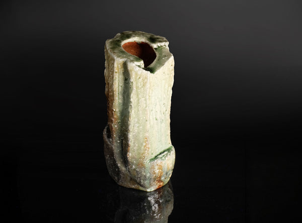 A rustic ceramic vase with a textured, weathered finish in off-white, green, and brown tones, featuring a small opening at the top, set against a dark reflective background.
