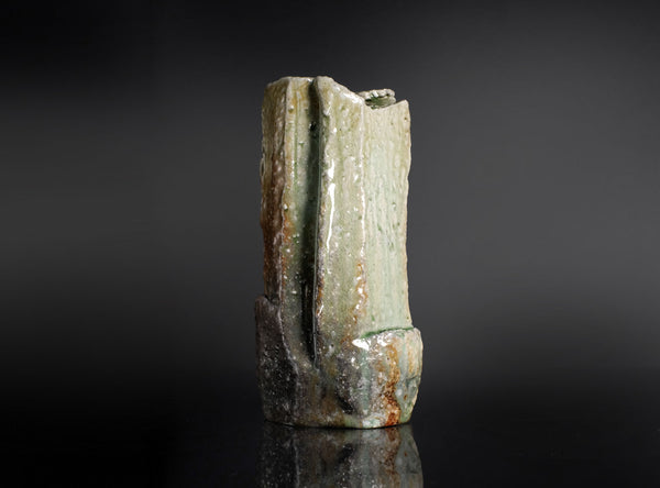 A handmade ceramic vase with a textured, rustic finish, featuring a vertical crack design and a natural earthy glaze in muted green, brown, and gray tones, set against a dark reflective background.