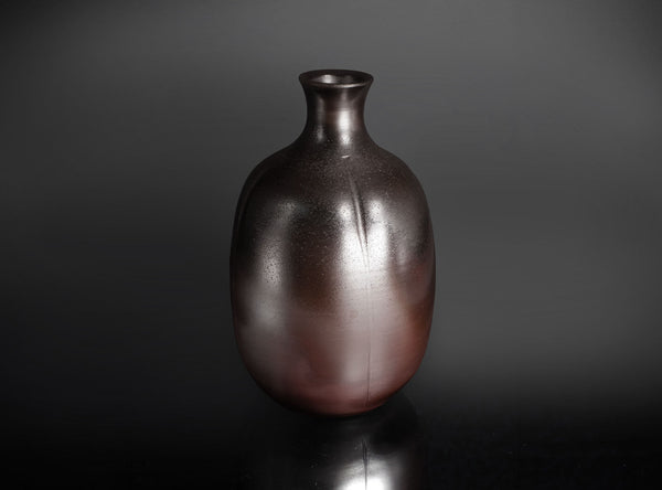 A dark brown ceramic bottle vase with a glossy, metallic finish, standing on a reflective black surface against a dark gray background.