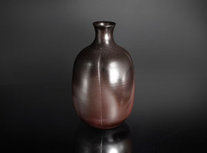 A small ceramic vase with a narrow neck and rounded body, featuring a metallic bronze to dark brown glaze with a subtle vertical seam, set against a dark gray gradient background with a soft reflection below.