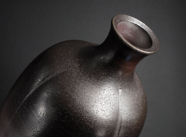 A close-up studio shot of a dark ceramic vase with a textured, matte finish and a flared neck, set against a dark gray background.