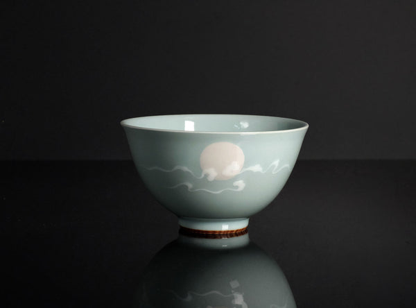 A pale celadon-green ceramic bowl with a hand-painted white moon and cloud motif, set against a dark reflective background.