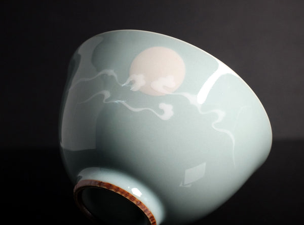A close-up of a glossy ceramic bowl with a pale celadon-green glaze, featuring a hand-painted white moon and cloud motif, set against a dark background.