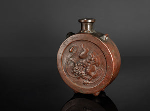 An antique bronze vase with a circular relief carving of a dragon on the front, set against a dark gray background.