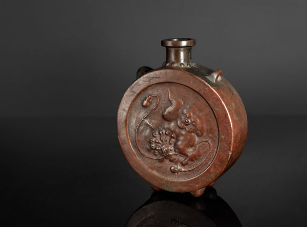 An antique bronze vase with a circular relief carving of a dragon on the front, set against a dark gray background.