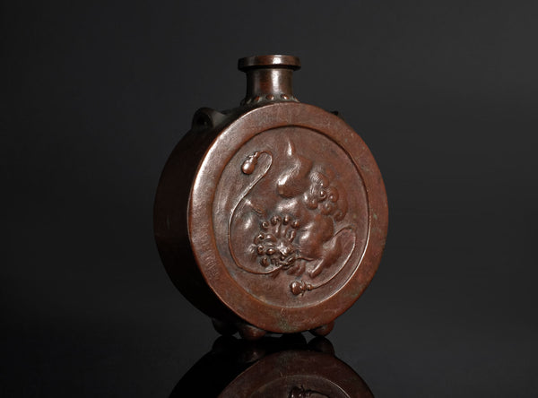 An antique bronze vase with a circular relief carving of a dragon on the front, set against a dark gray background.
