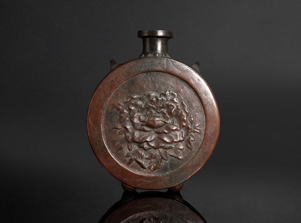 An antique round copper vase with a floral relief design on the front, set against a dark gray background.