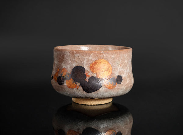 A handmade ceramic tea bowl with a textured, earthy brown and beige glaze, decorated with abstract hand-painted black and orange spots, set against a dark reflective background.