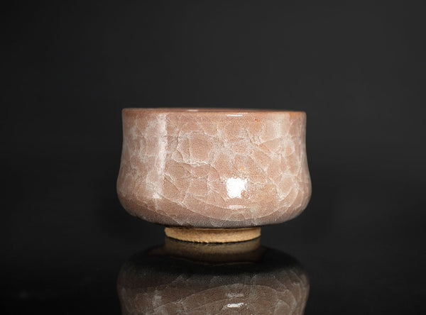 A handmade ceramic bowl with a textured, crackled glaze in a dusty rose pink color, resting on a reflective black surface against a dark background.