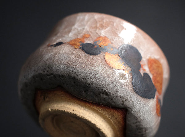 A close-up of a handmade ceramic bowl with a textured, speckled gray and brown glaze, accented with organic black and rust-orange patches, set against a dark background.