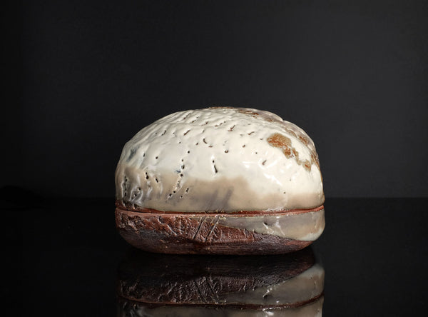A handmade ceramic lidded box with a textured, organic-shaped cream and brown glazed top and a rustic dark brown base, displayed on a reflective black surface against a black background.
