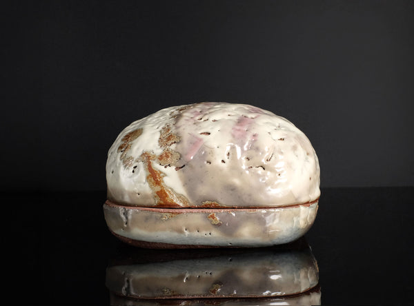 A handmade ceramic lidded box with a textured, organic-shaped cream and brown glazed top and a rustic dark brown base, displayed on a reflective black surface against a black background.
