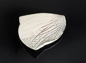 A ceramic leaf-shaped dish or plate with a glossy white glaze and textured crackled detailing, displayed on a reflective black surface against a dark background.