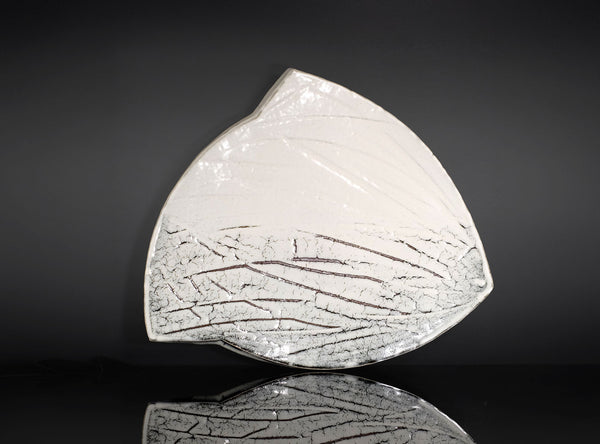 An organic-shaped white ceramic plate with a textured, crackled gray pattern on the bottom half, displayed on a reflective black surface against a dark gray background.